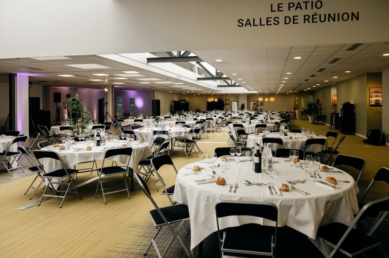 Salle de réception lumineuse avec tables rondes dressées pour un événement, chaises noires et décoration sobre.