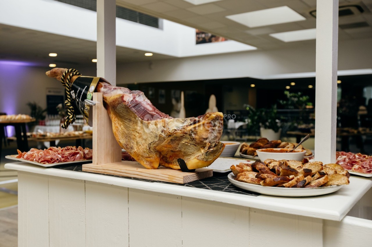 Jambon entier sur support en bois, entouré d'assiettes de charcuterie et de pain grillé dans un espace lumineux.