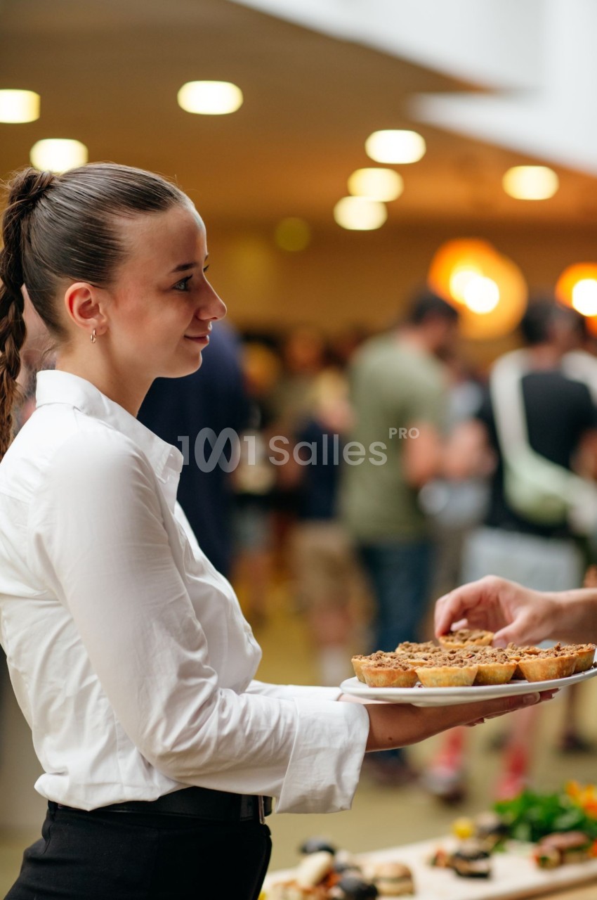 Une serveuse en chemise blanche tend un plat de desserts dans un espace intérieur animé.