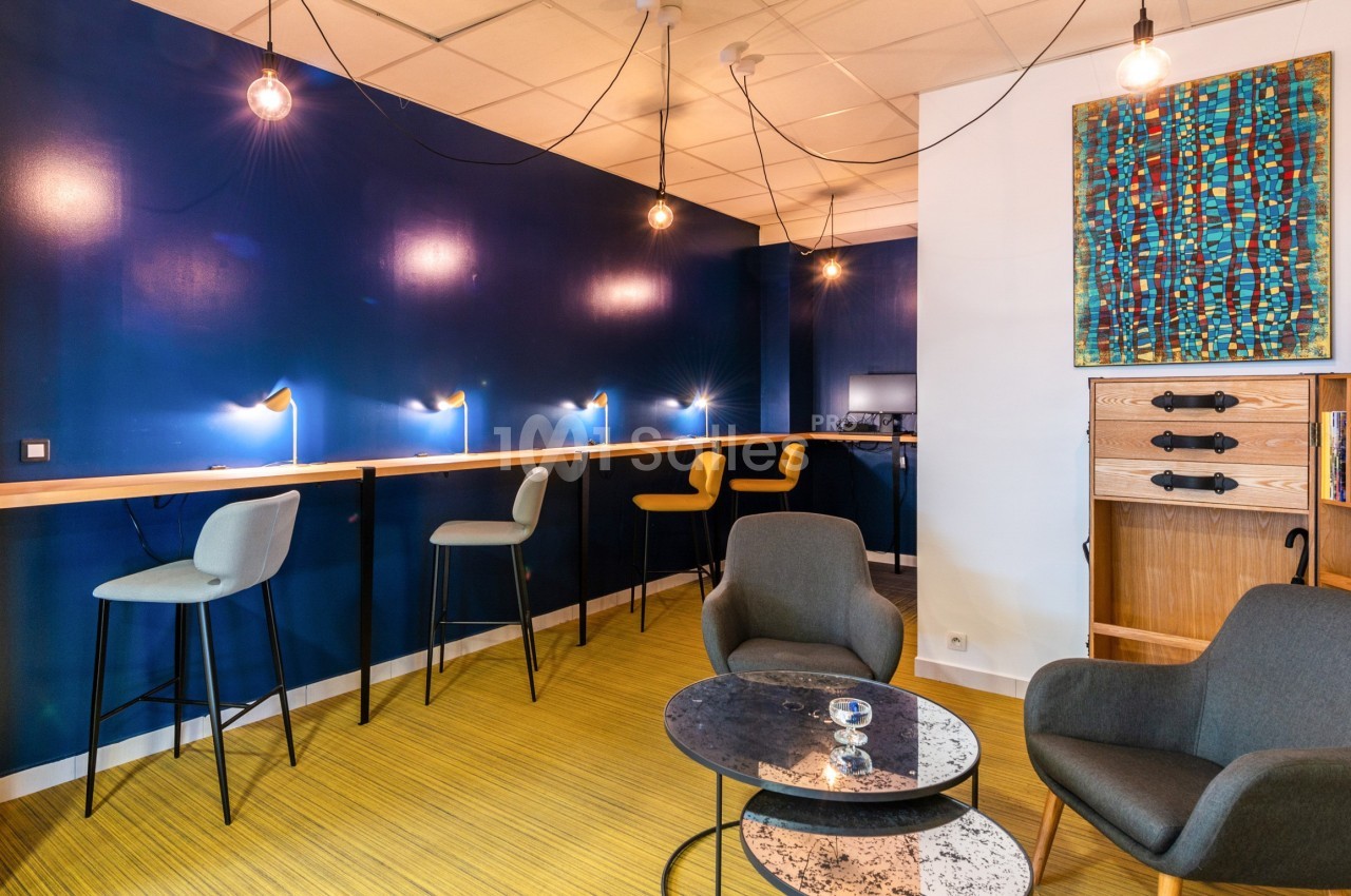 Espace de travail moderne avec murs bleus, chaises hautes, fauteuils, table basse et éclairage suspendu.