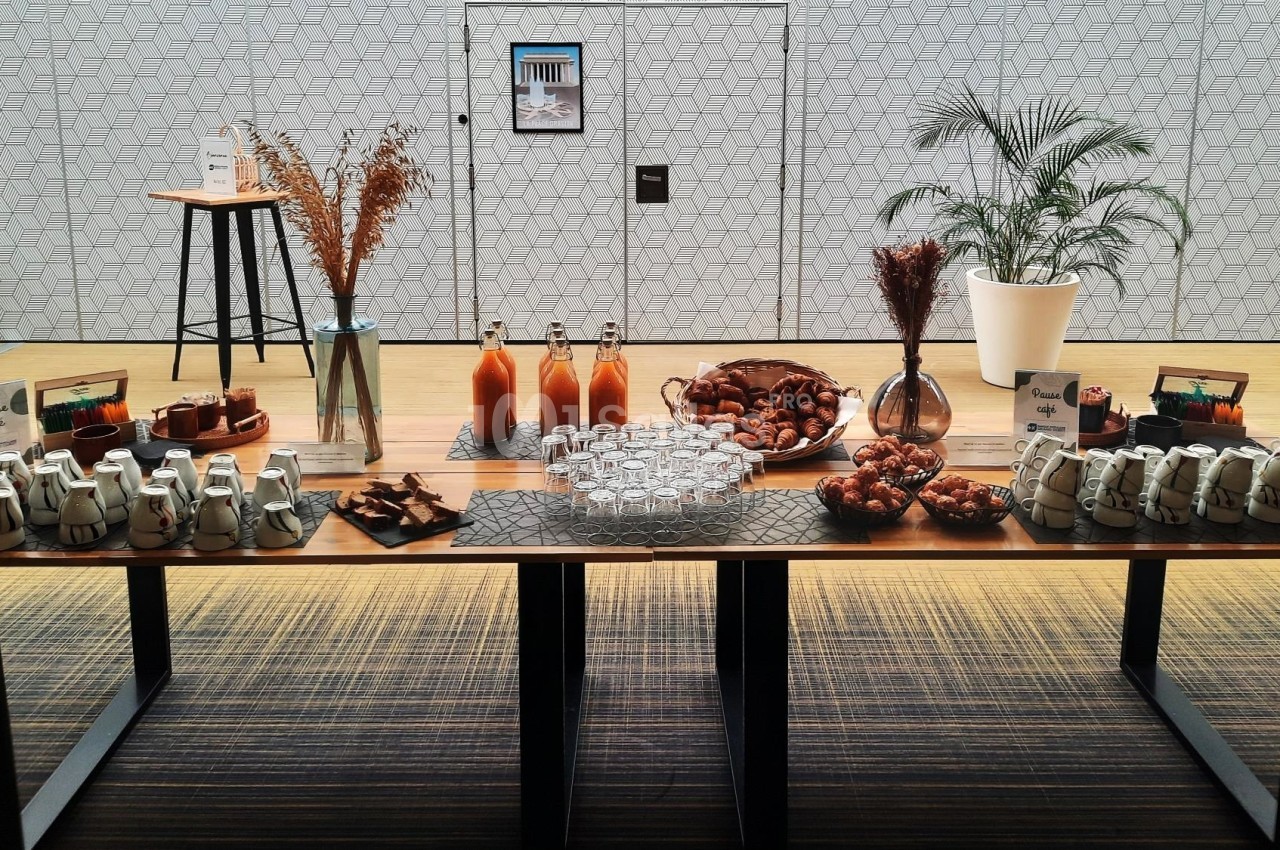 Buffet composé de boissons, viennoiseries, pots de yaourt et décorations sur une table en bois dans une salle lumineuse.