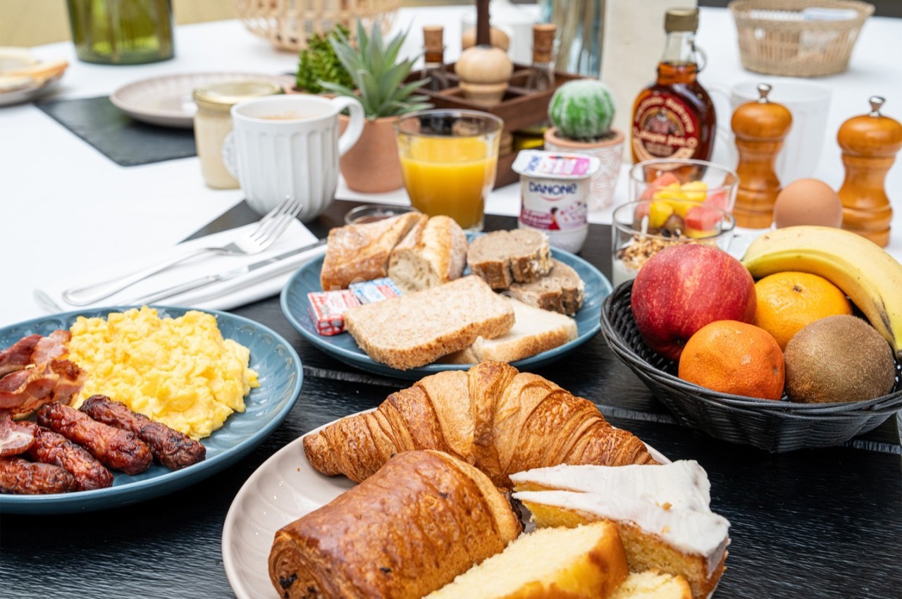 Petit-déjeuner varié avec viennoiseries, fruits, pain, confitures, œufs brouillés, saucisses et boissons.