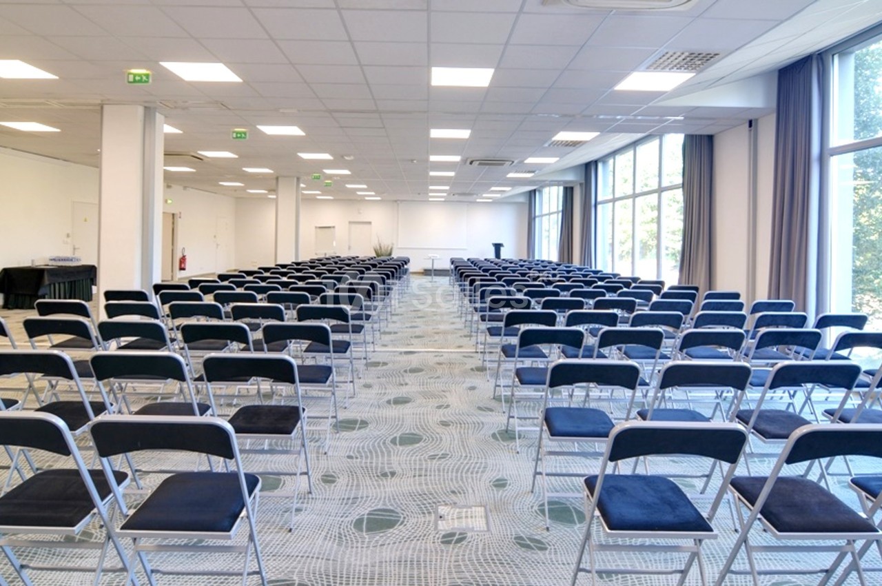 Salle de conférence lumineuse avec rangées de chaises pliantes noires alignées face à un pupitre.