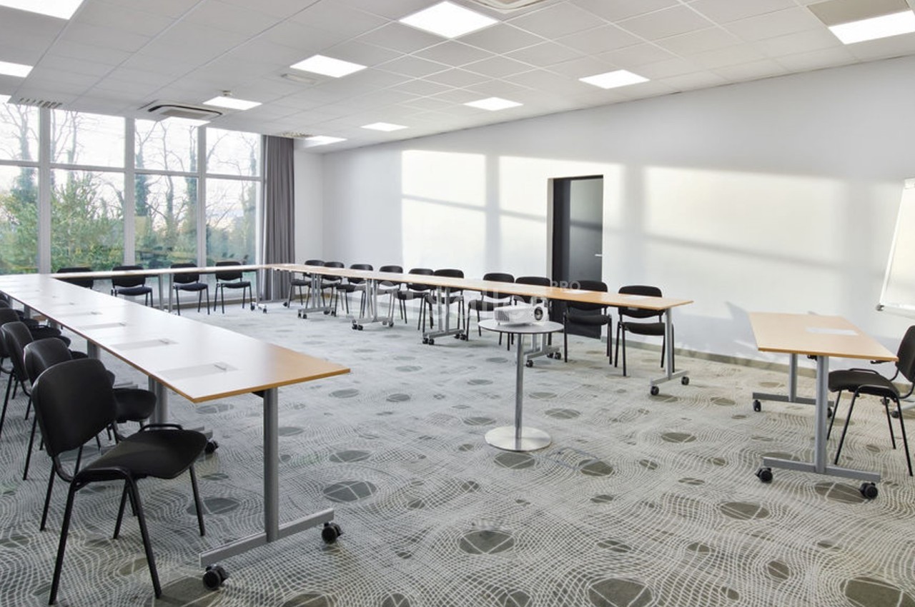 Salle de réunion lumineuse avec tables disposées en U, chaises noires, tableau blanc et projecteur.