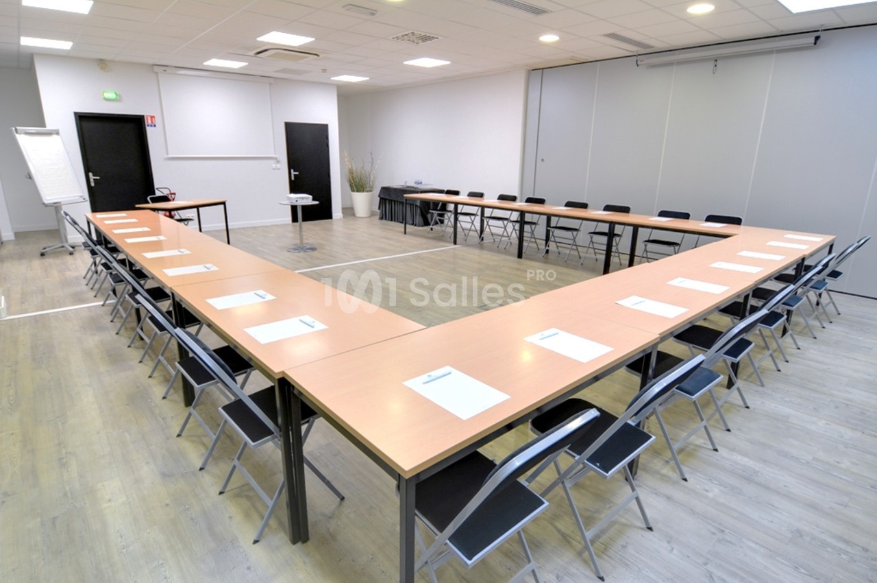 Salle de réunion lumineuse avec tables disposées en U, chaises pliantes et paperboard dans un coin.