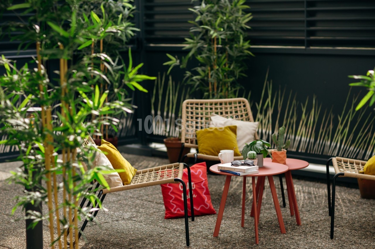 Deux fauteuils en rotin avec coussins colorés entourent une table basse rouge sur une terrasse végétalisée.