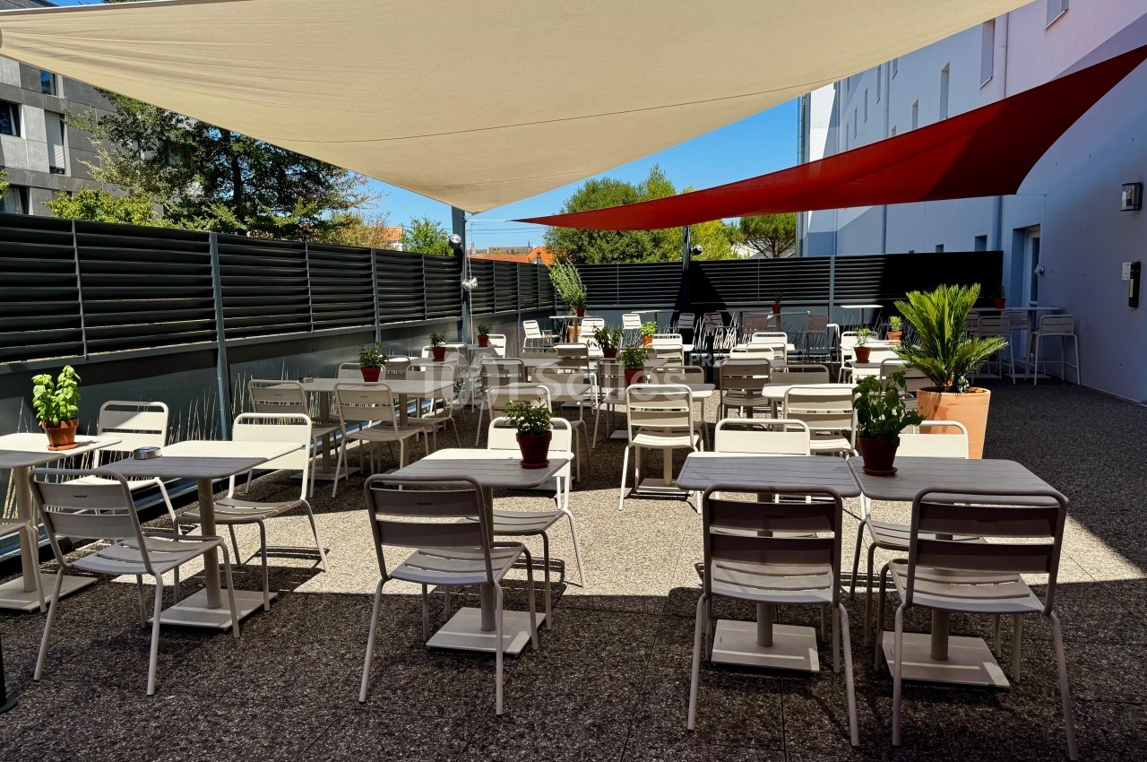 Terrasse extérieure avec tables et chaises blanches, ombragée par des voiles tendues, entourée de plantes et de panneaux.