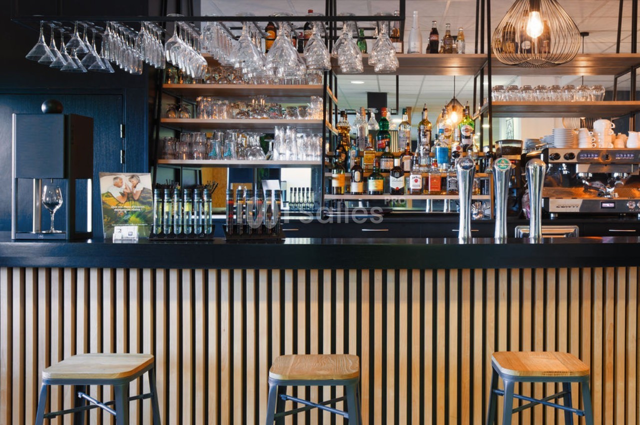 Comptoir de bar moderne avec tabourets en bois, étagères garnies de bouteilles et verres suspendus au-dessus.