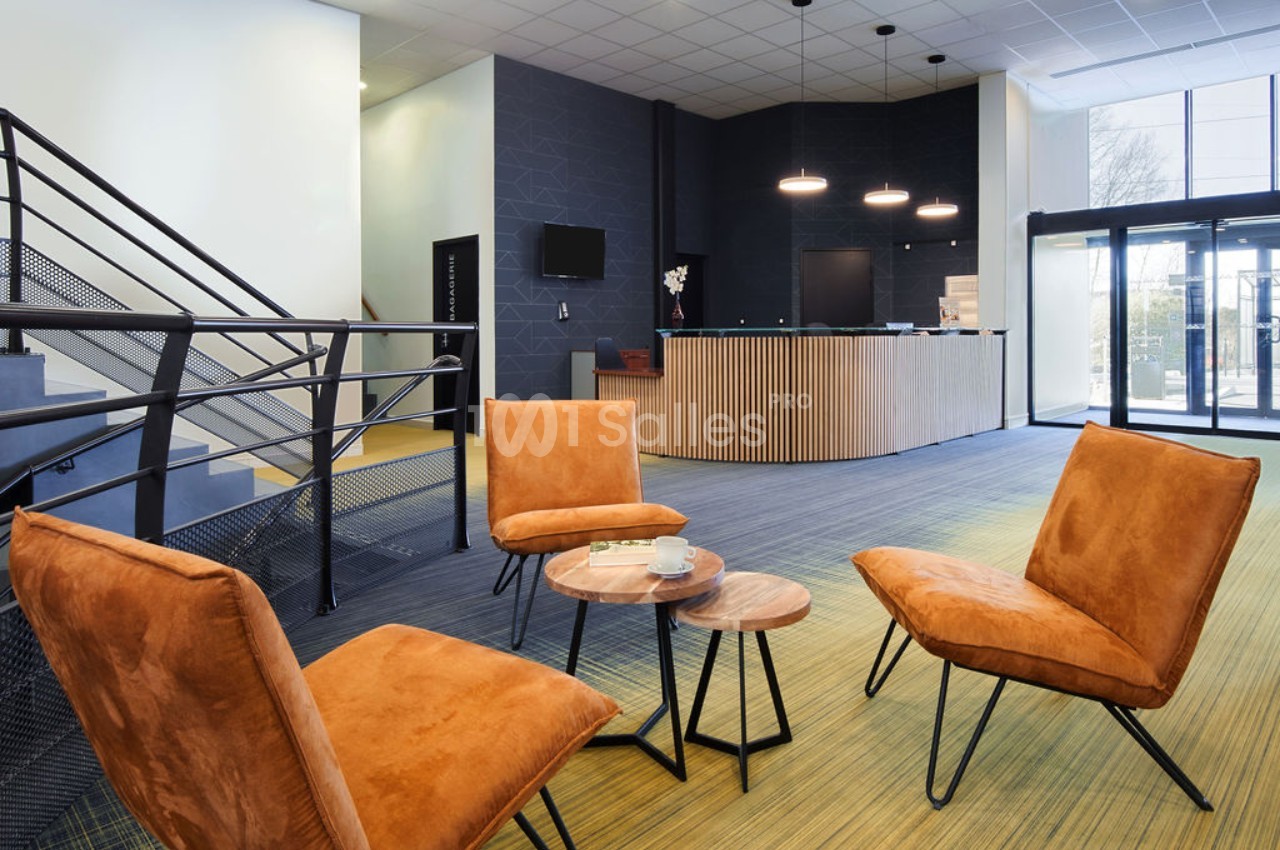 Espace d'accueil moderne avec fauteuils orange, tables basses en bois, escalier métallique et comptoir de réception.