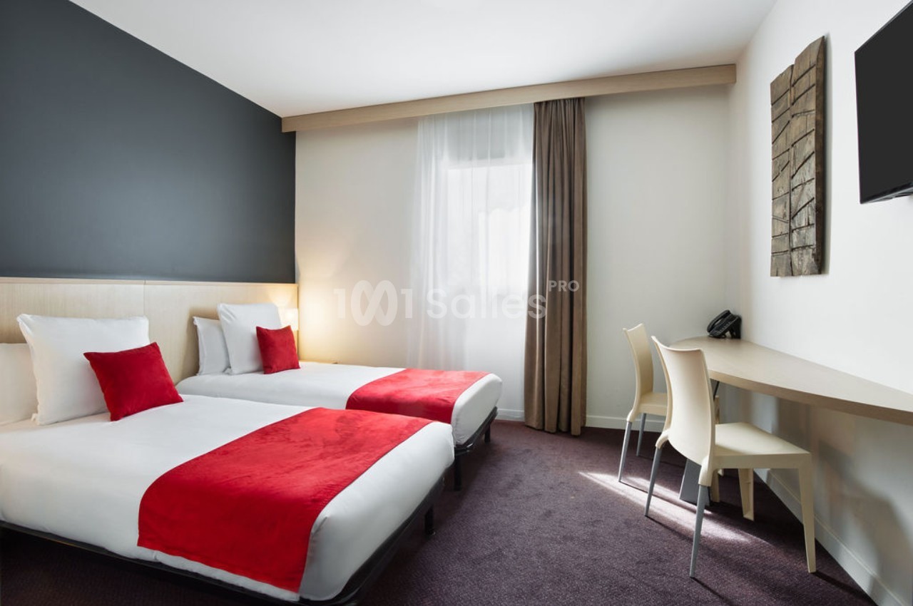 Chambre d'hôtel moderne avec deux lits simples, couvre-lits rouges, bureau, chaises et grande fenêtre avec rideaux.