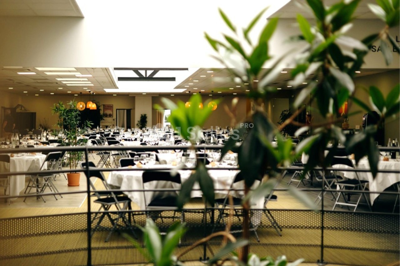 Salle de réception lumineuse avec des tables rondes dressées, entourées de chaises noires, et des plantes au premier plan.