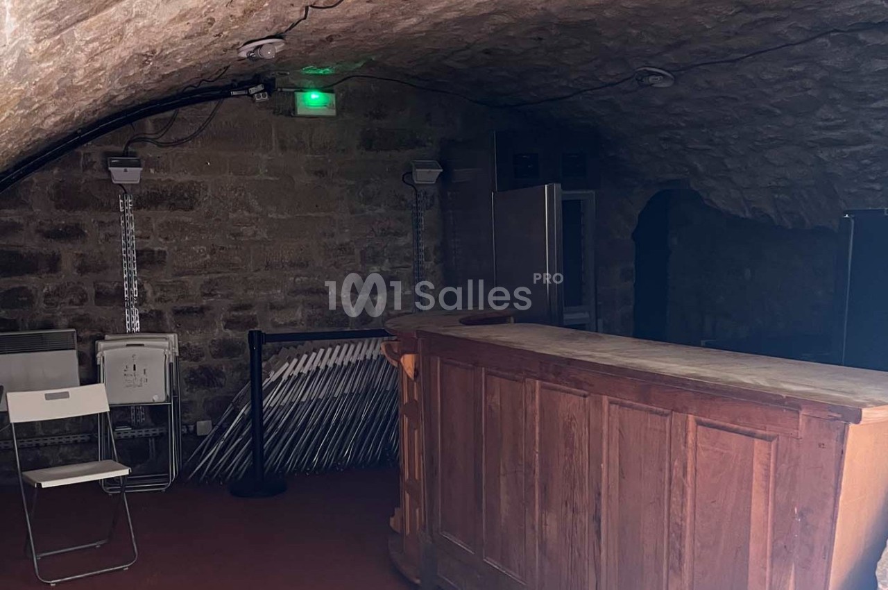 Cave voûtée en pierre avec un comptoir en bois, des chaises pliées et un éclairage tamisé.