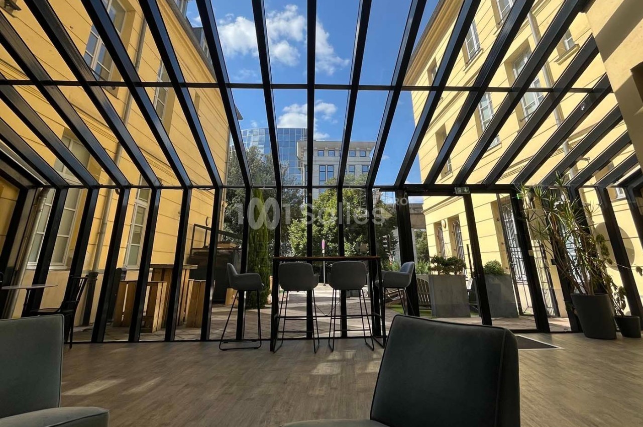 Espace intérieur lumineux avec verrière, mobilier moderne et vue sur une cour arborée sous un ciel bleu.