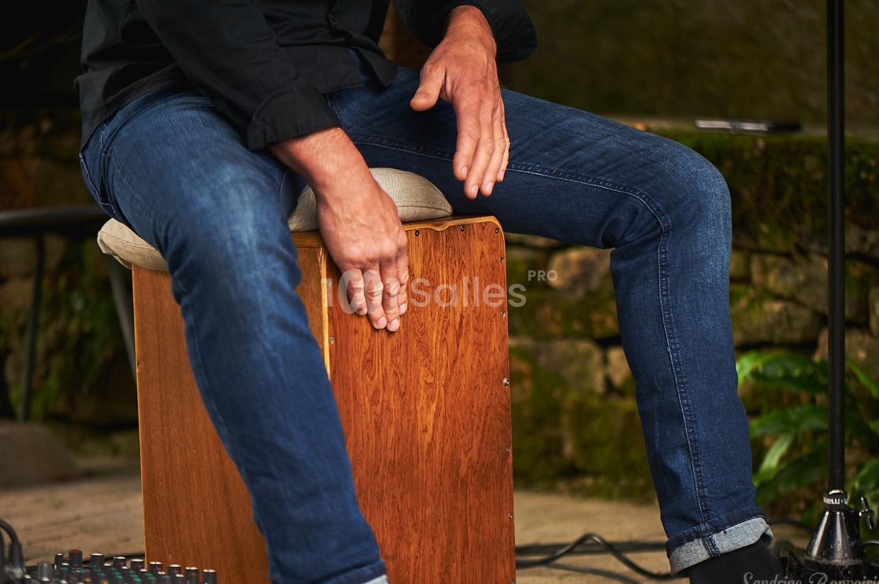 Musicien jouant du cajón en bois, ses mains frappant le devant de l'instrument dans un cadre extérieur.