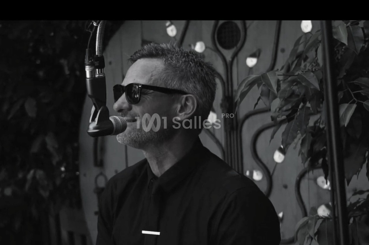 Homme portant des lunettes de soleil, chantant au microphone devant un décor végétal et artistique.