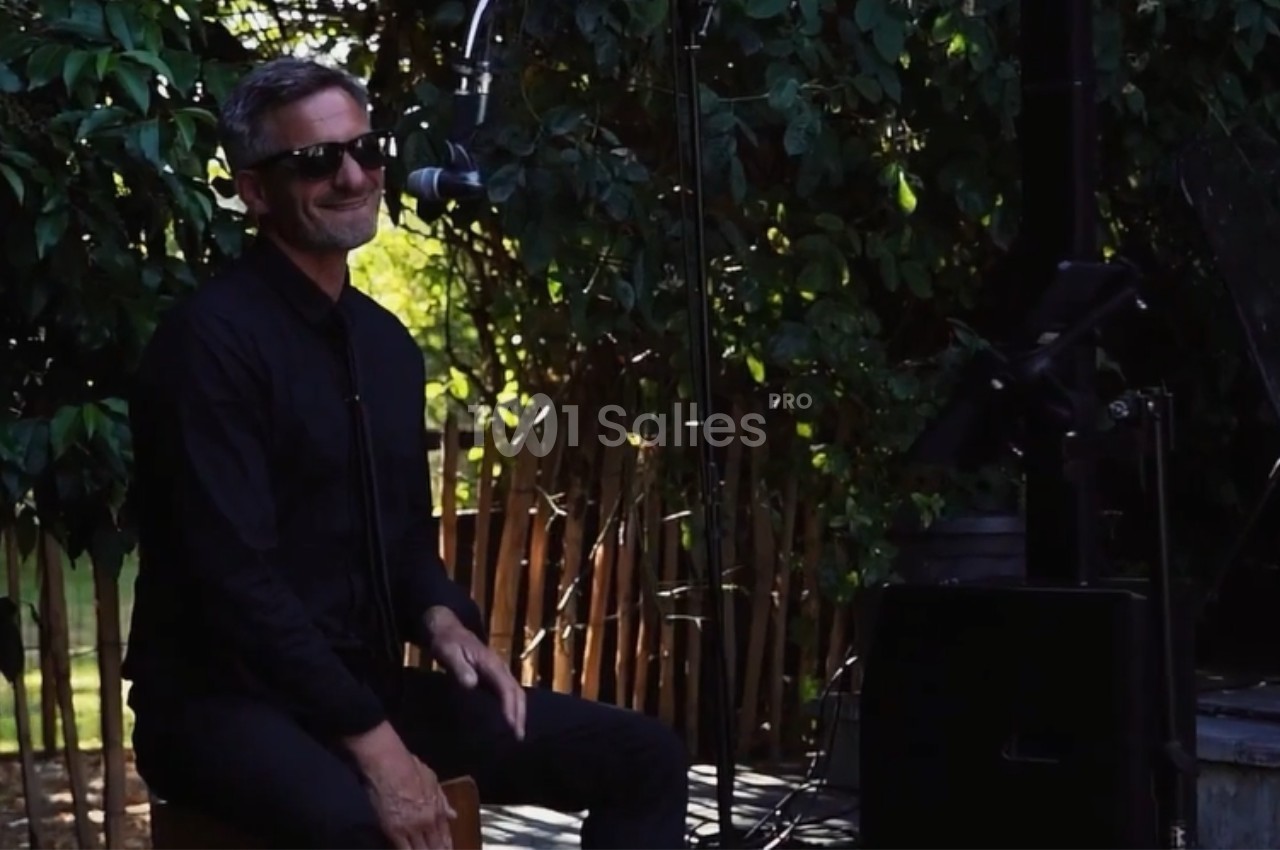 Un musicien souriant joue du cajón en extérieur, entouré de verdure et équipé d'un micro.