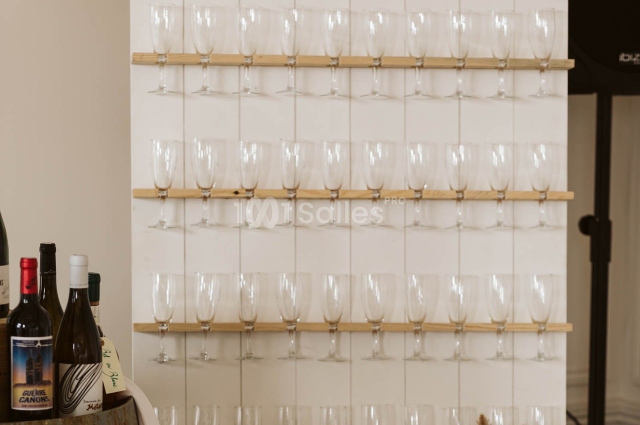 Des verres à vin vides alignés sur des étagères en bois fixées à un mur blanc, avec des bouteilles au premier plan.