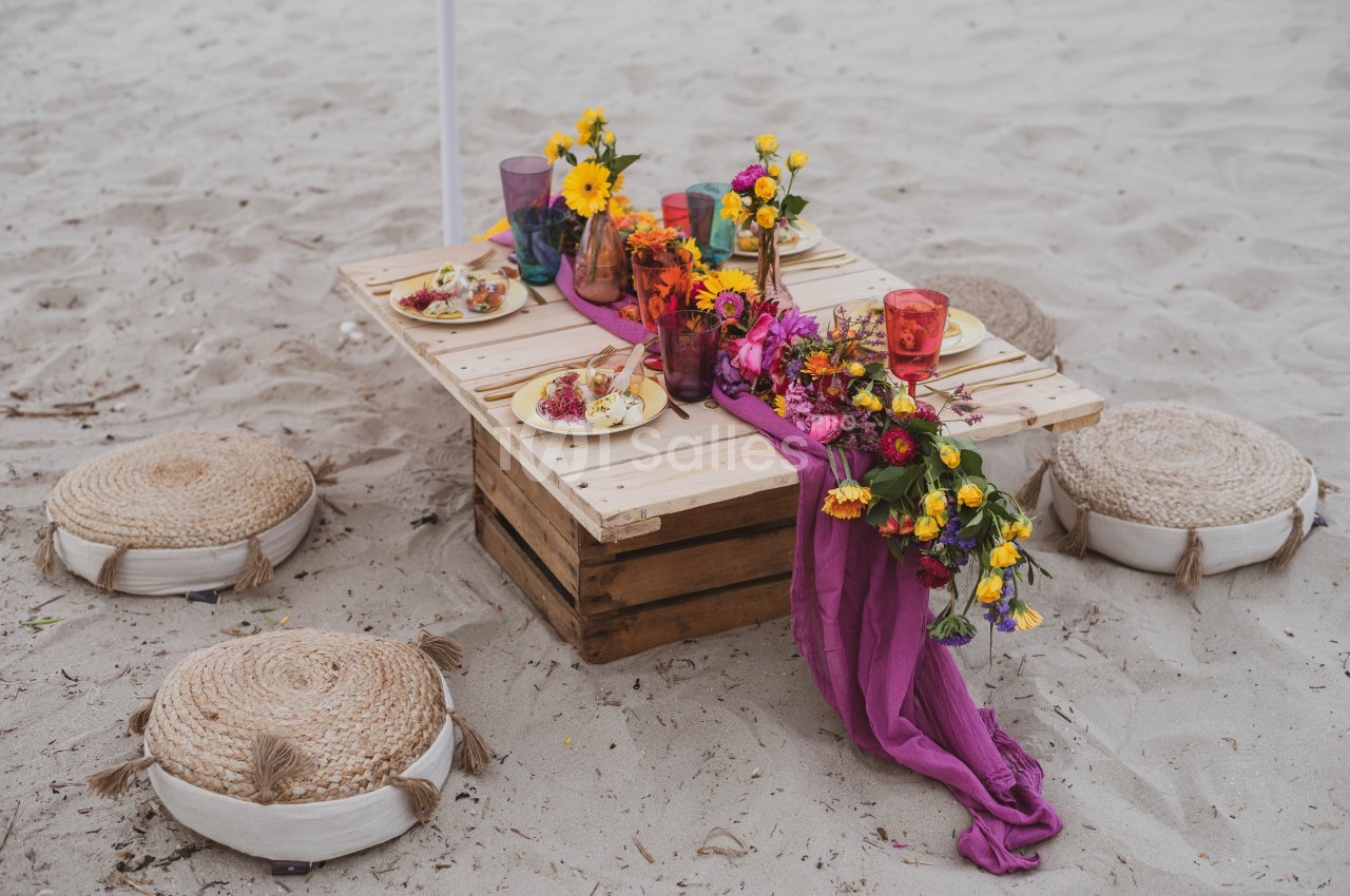 Table basse en bois décorée de fleurs colorées, vaisselle et tissu violet, entourée de coussins sur du sable.