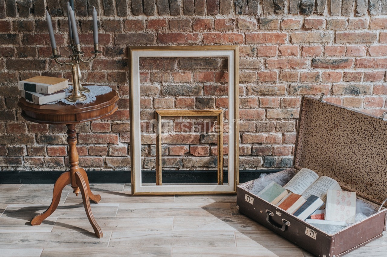 Table en bois avec chandelier et livres, cadres dorés appuyés contre un mur de briques, valise ouverte contenant des…