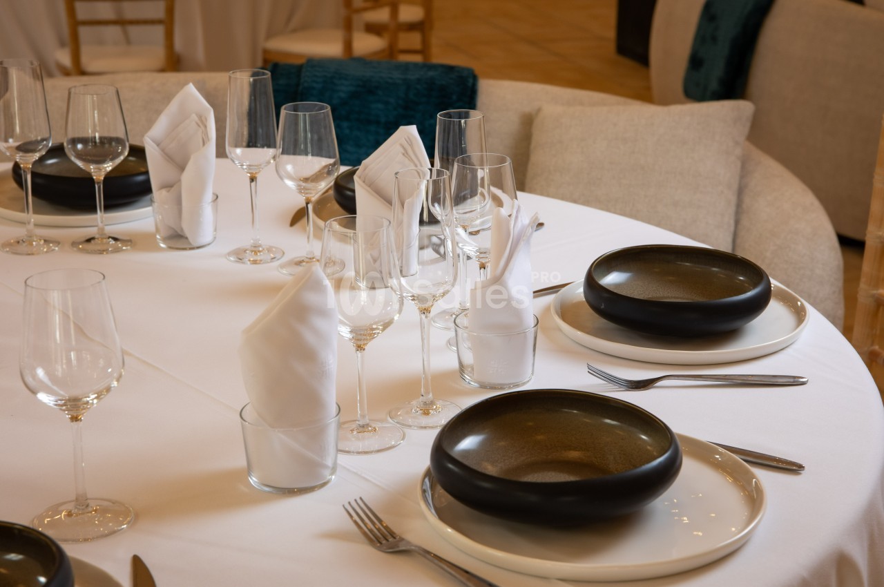 Table dressée avec nappes blanches, assiettes noires, verres à pied et serviettes pliées.