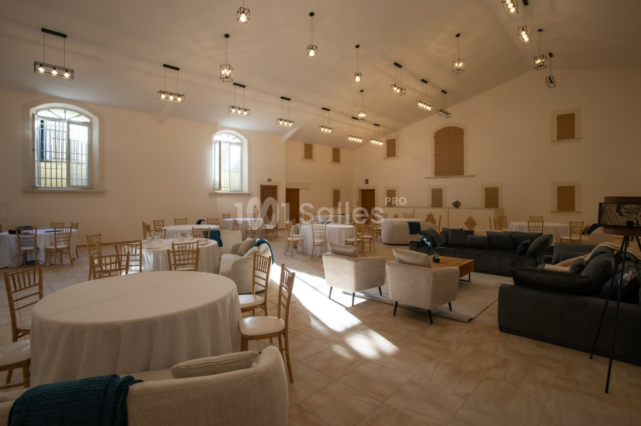 Salle lumineuse avec tables rondes dressées, chaises en bois, canapés et grandes fenêtres laissant entrer la lumière…