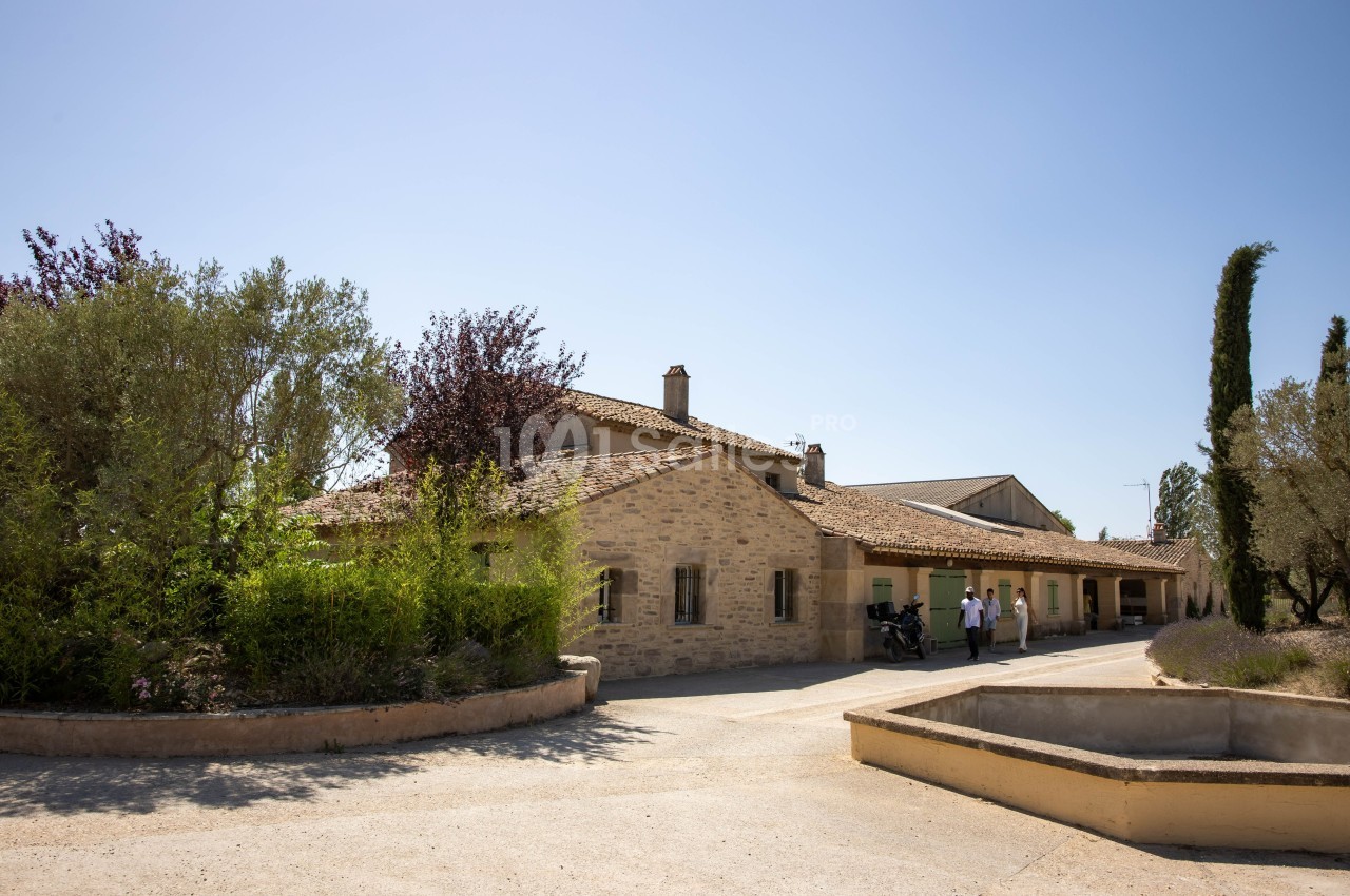 Bâtiment en pierre entouré de végétation méditerranéenne sous un ciel dégagé, avec quelques personnes à l'entrée.
