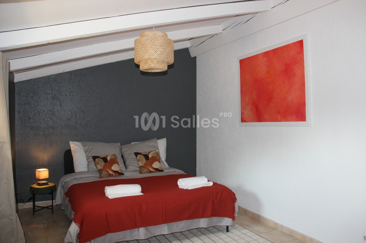 Chambre mansardée avec lit double, couvre-lit rouge, mur gris, tableau abstrait rouge et lampe suspendue en osier.