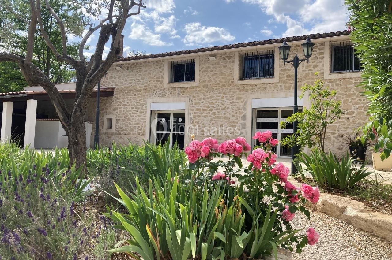 Façade d'une maison en pierre entourée de végétation, avec des fleurs roses au premier plan et un ciel partiellement nuageux.