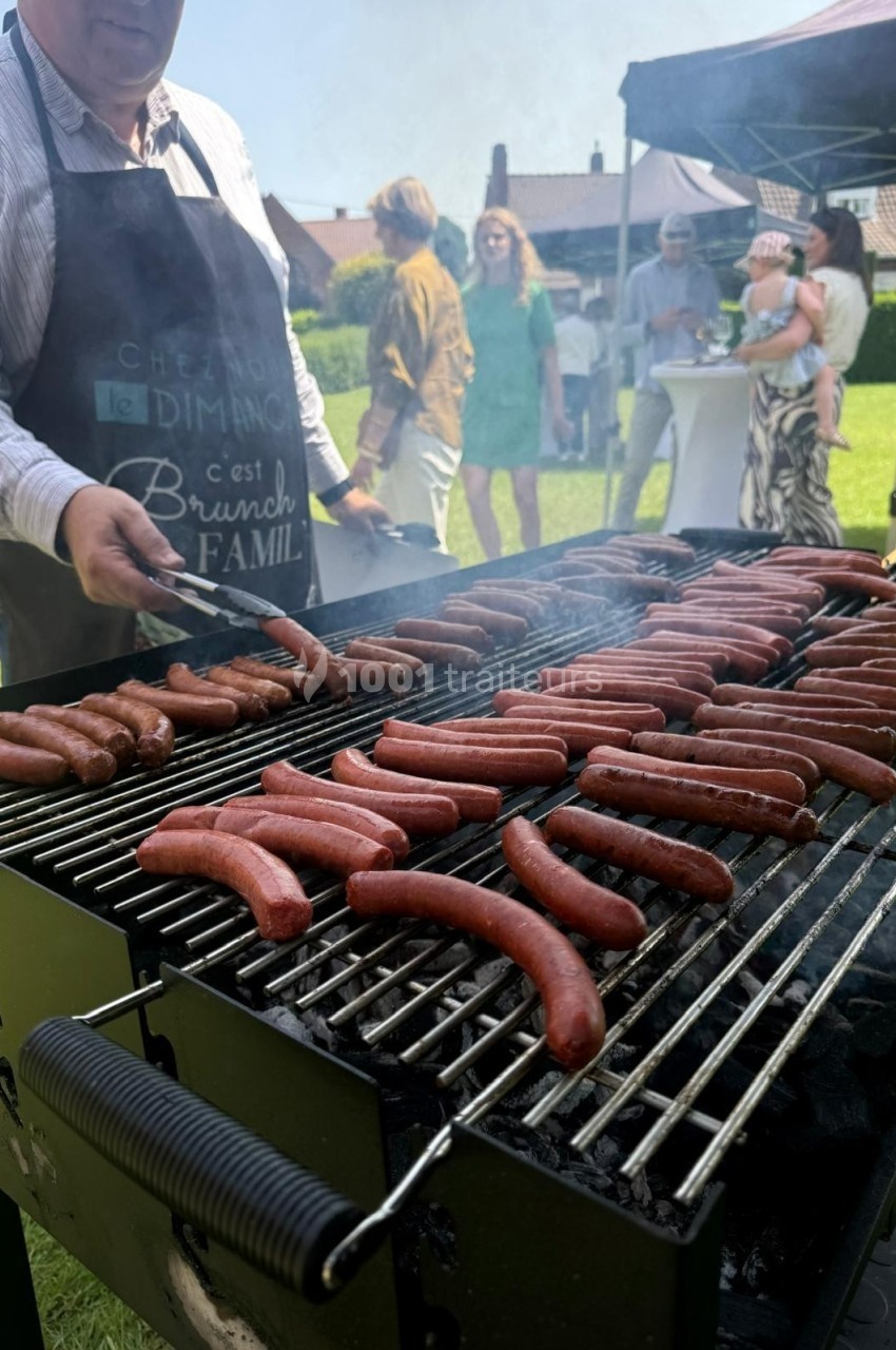 Un homme en tablier cuisine des saucisses sur un grand barbecue lors d'un événement en plein air.