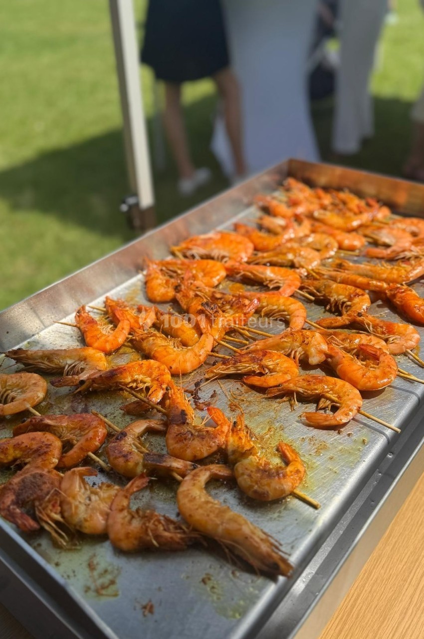 Crevettes grillées sur une plaque de cuisson en extérieur, avec des personnes floues en arrière-plan.