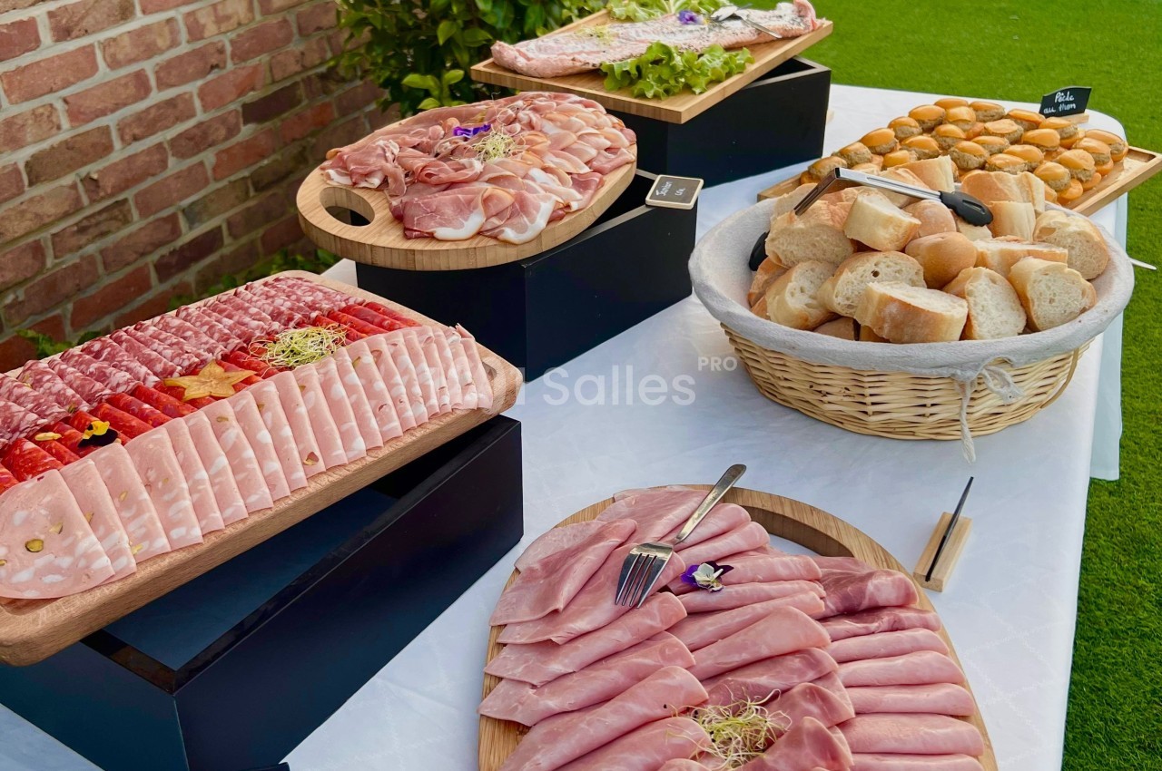 Plateau de charcuterie variée, pain et petits pains disposés sur une table en extérieur avec nappe blanche.