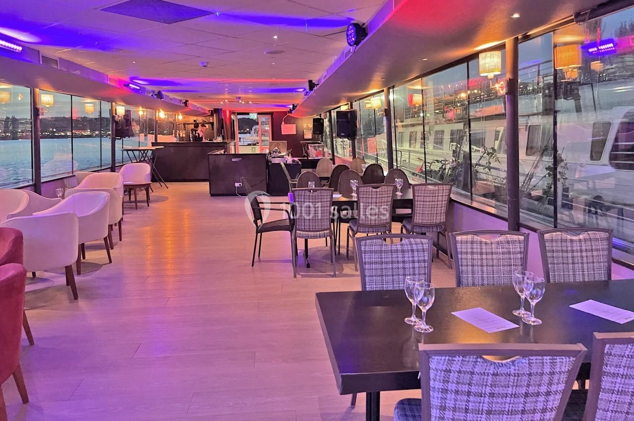 Salle de restaurant lumineuse avec grandes baies vitrées donnant sur l'extérieur, tables dressées et éclairage chaleureux.