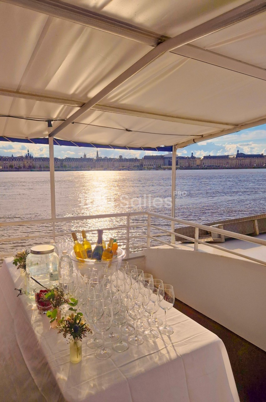 Table dressée avec des verres, carafes et fleurs, sur un bateau avec vue sur un fleuve et une ville en arrière-plan.