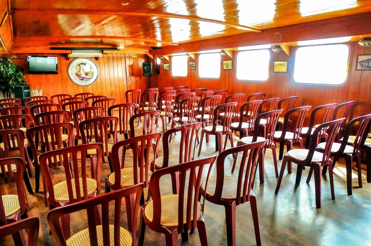 Salle en bois avec des rangées de chaises vides en plastique et assises en osier, éclairée par des fenêtres latérales.