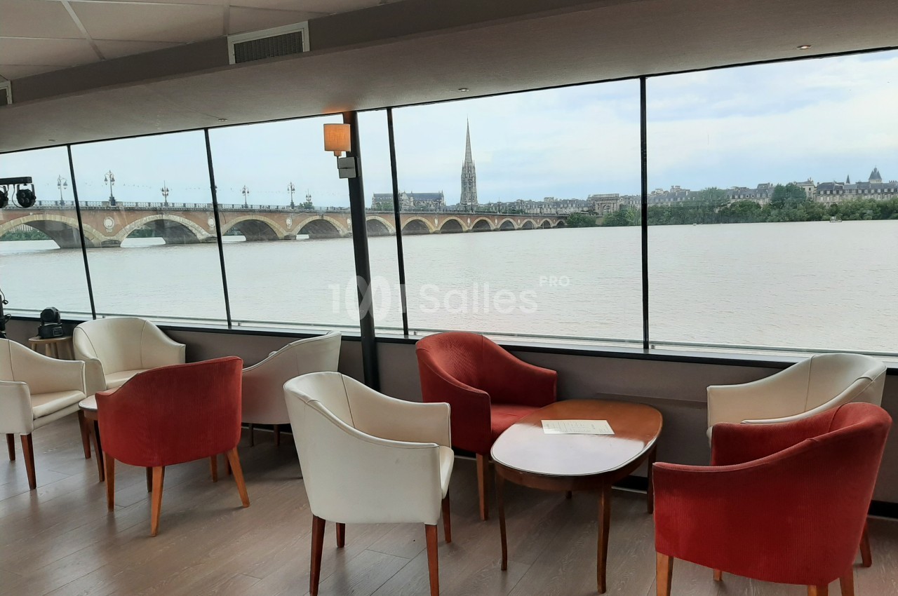 Salon avec fauteuils rouges et blancs, tables en bois, et vue sur un fleuve, un pont et une église à l'extérieur.