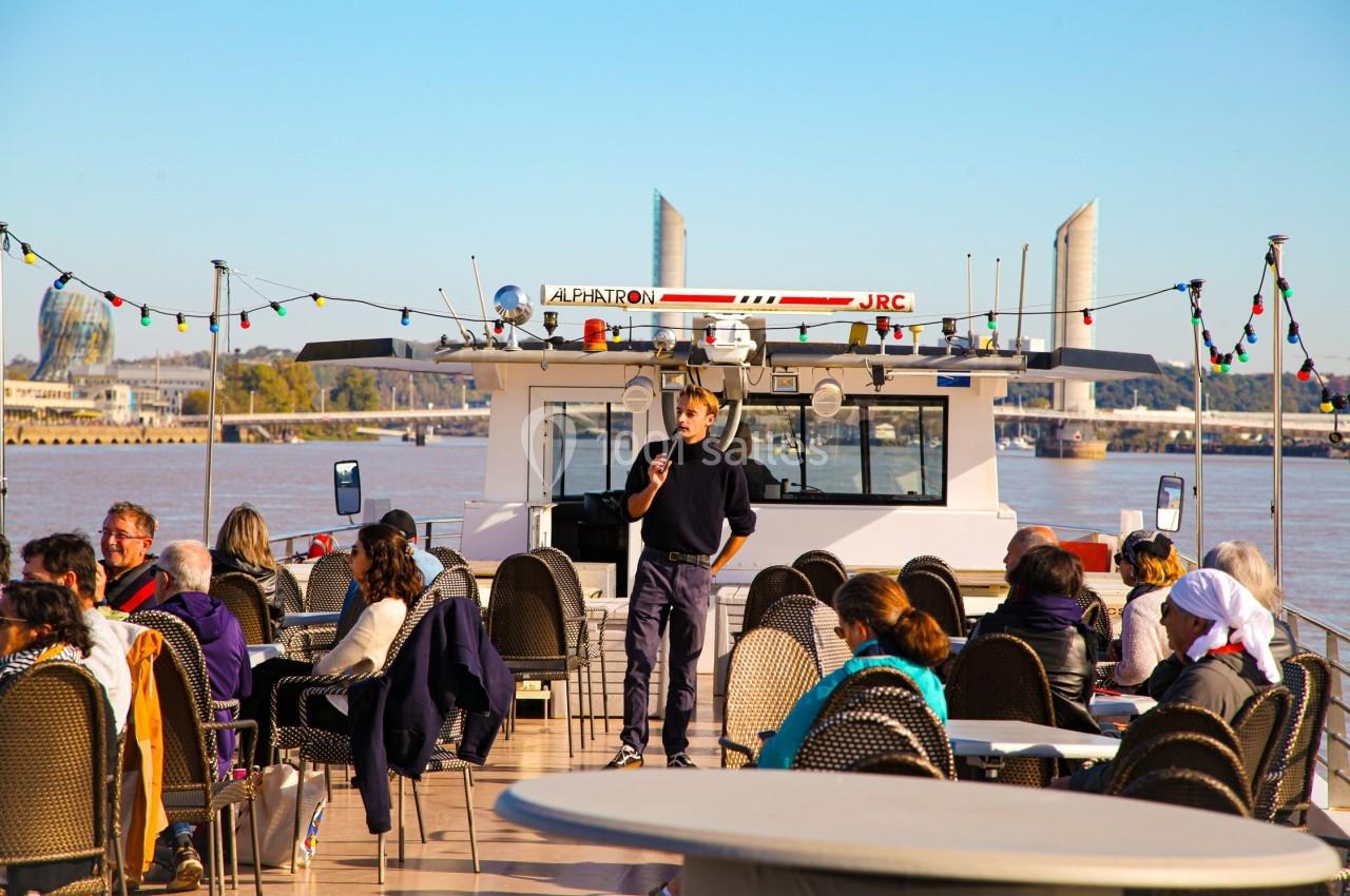 Un guide s'adresse à un groupe de personnes assises sur le pont d'un bateau naviguant sur un fleuve, par temps clair.