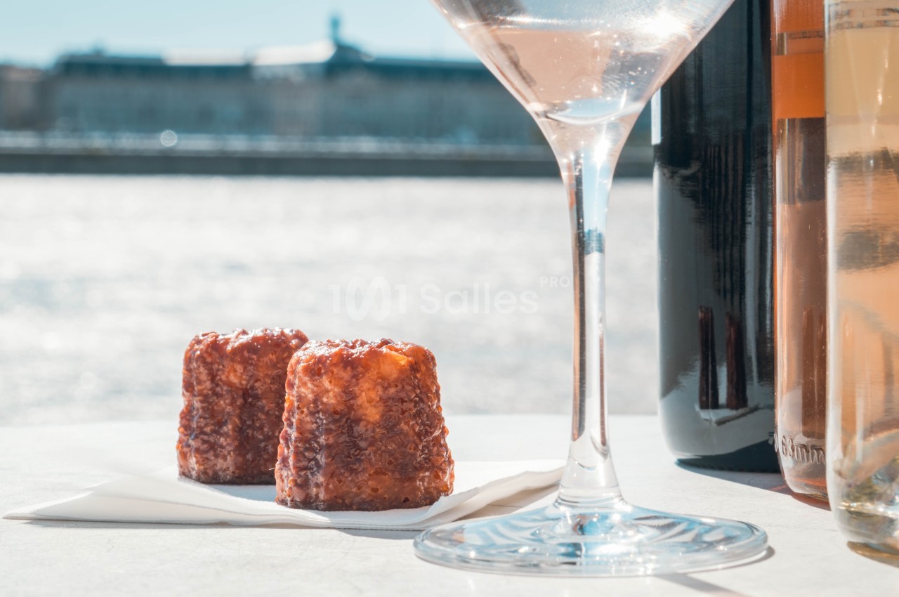 Deux cannelés posés sur une serviette blanche, entourés de bouteilles de vin et d'un verre, avec un fond fluvial ensoleillé.