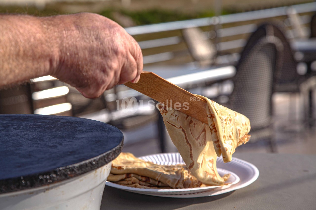 Une main dépose une crêpe chaude sur une assiette à l'aide d'une spatule en bois près d'une crêpière.