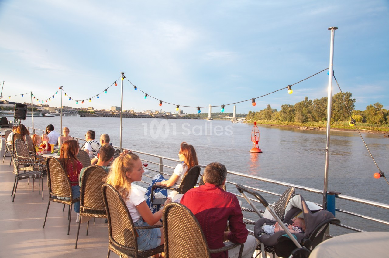 Des personnes assises sur le pont d'un bateau, profitant d'une vue sur une rivière bordée de végétation et de bouées.