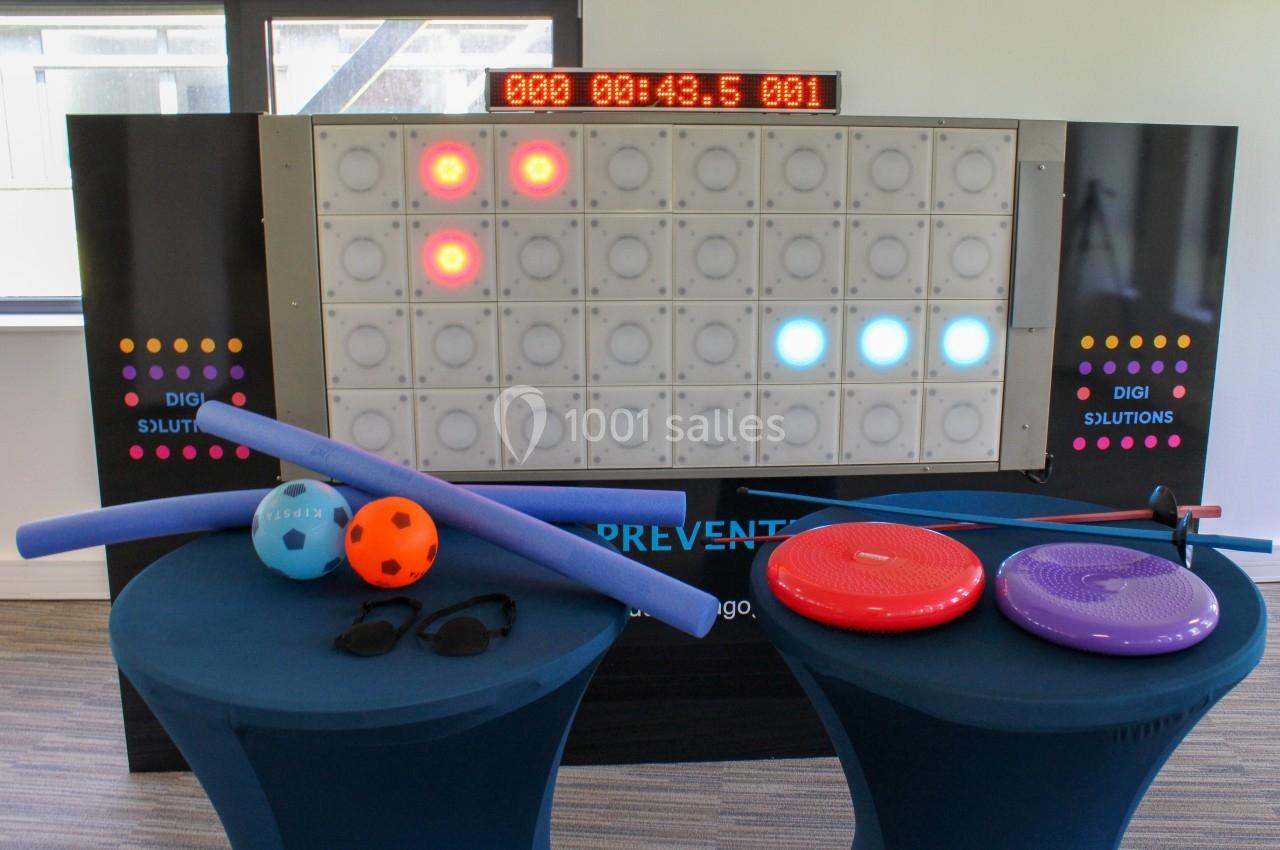 Mur interactif lumineux avec chronomètre, accessoires de sport et équilibre disposés sur deux tables bleues au premier plan.