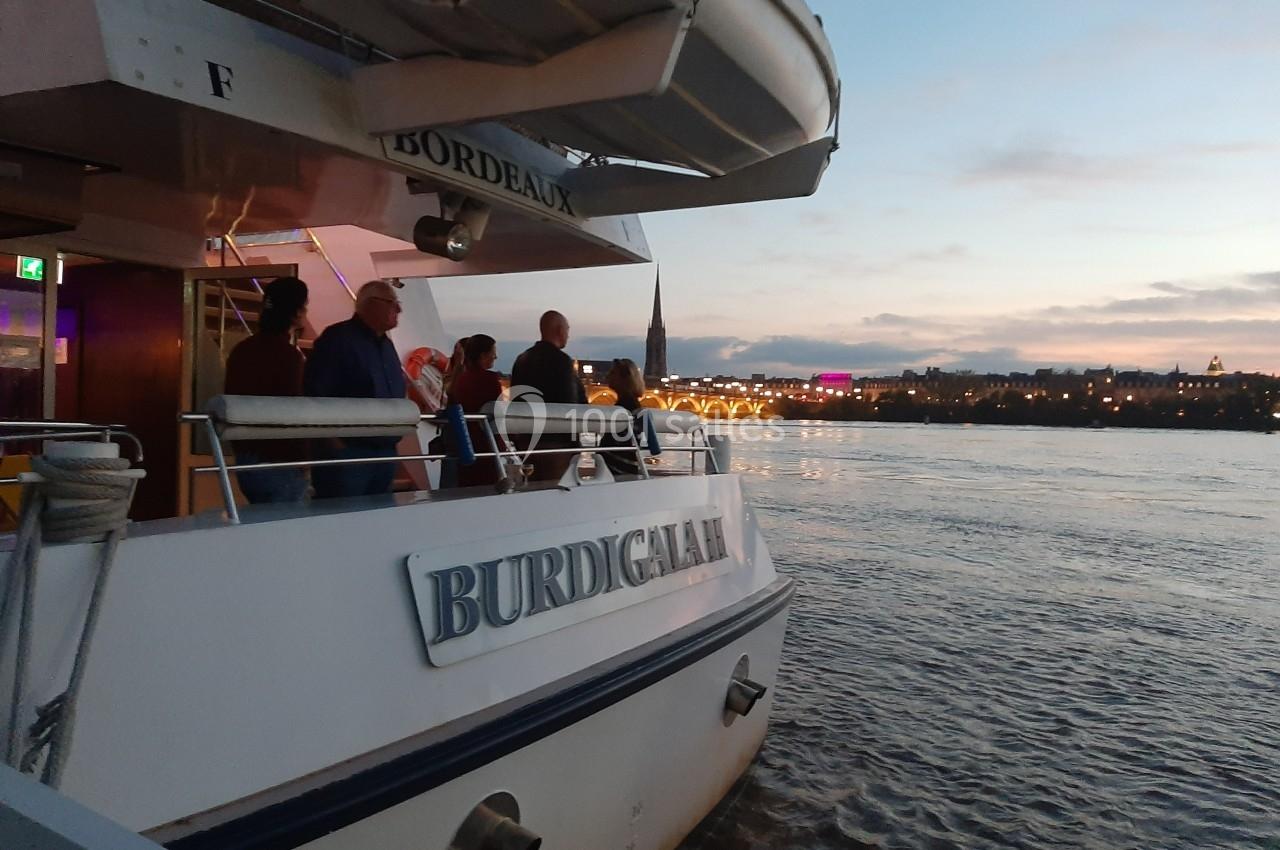 Des passagers sur le pont d'un bateau nommé ’Burdigala’, naviguant sur un fleuve au crépuscule avec une ville illuminée en…