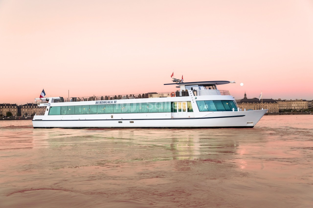 Bateau blanc naviguant sur une rivière au coucher du soleil, avec des bâtiments historiques en arrière-plan.