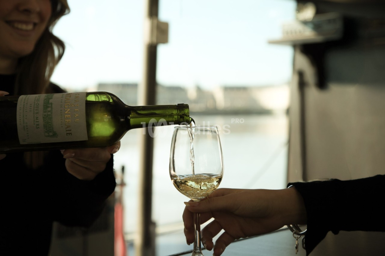 Une personne verse du vin blanc dans un verre tenu par une autre personne, en arrière-plan flou d'une rivière.