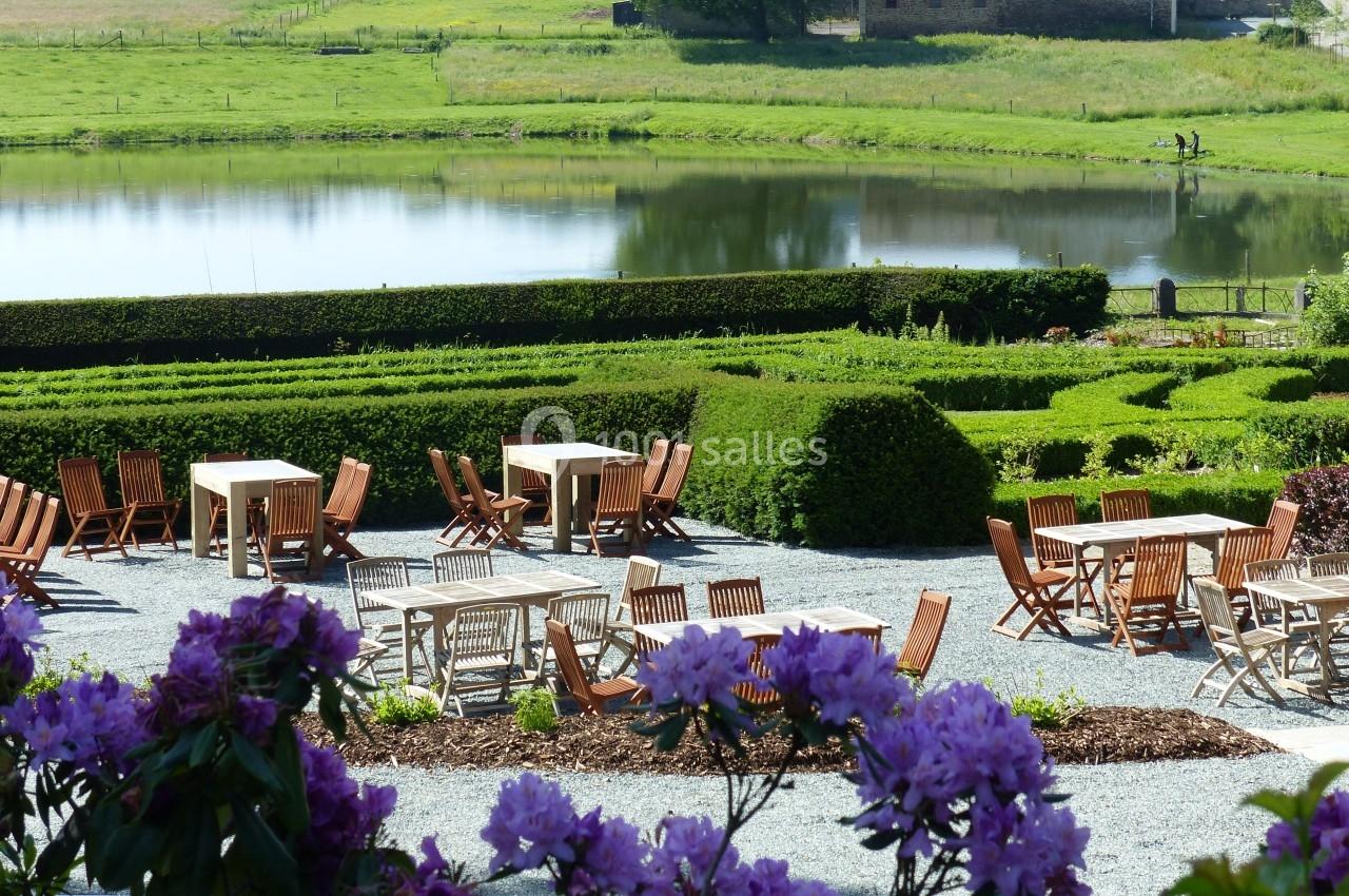 Terrasse avec tables et chaises en bois, entourée de buissons taillés, donnant sur un étang et un paysage verdoyant.