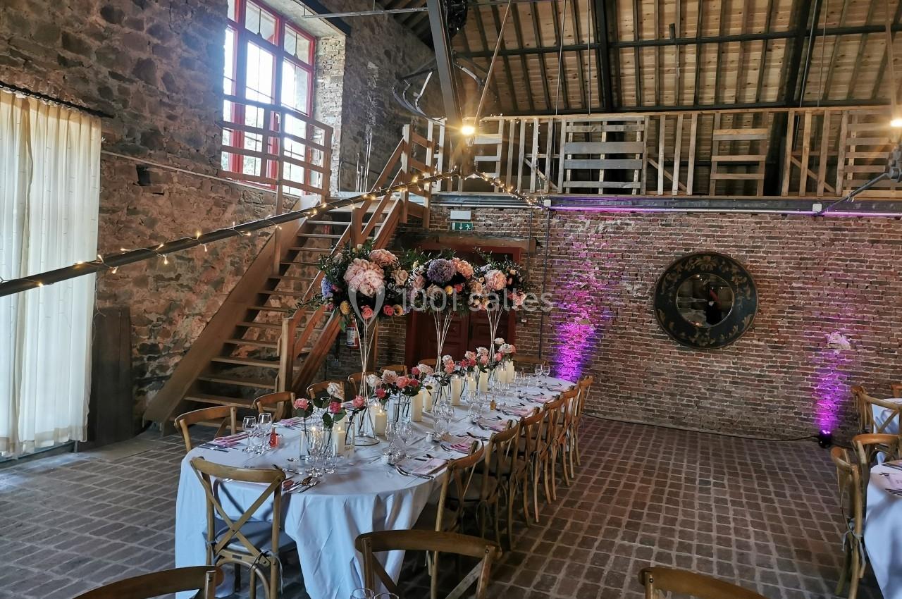 Salle de réception rustique avec tables décorées de fleurs, murs en briques, poutres apparentes et éclairage tamisé.