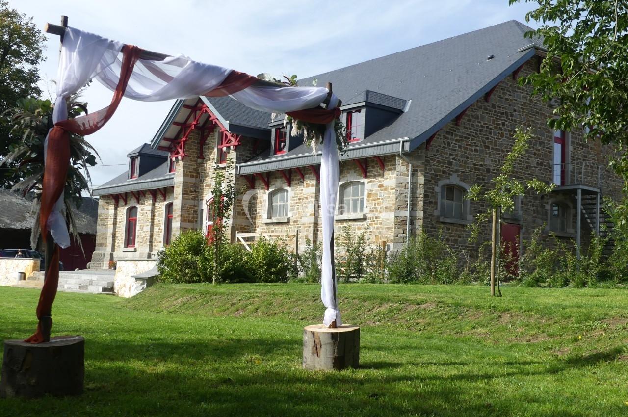 Structure en pierre avec toit en ardoise, vue depuis un jardin avec une arche décorée de tissu au premier plan.