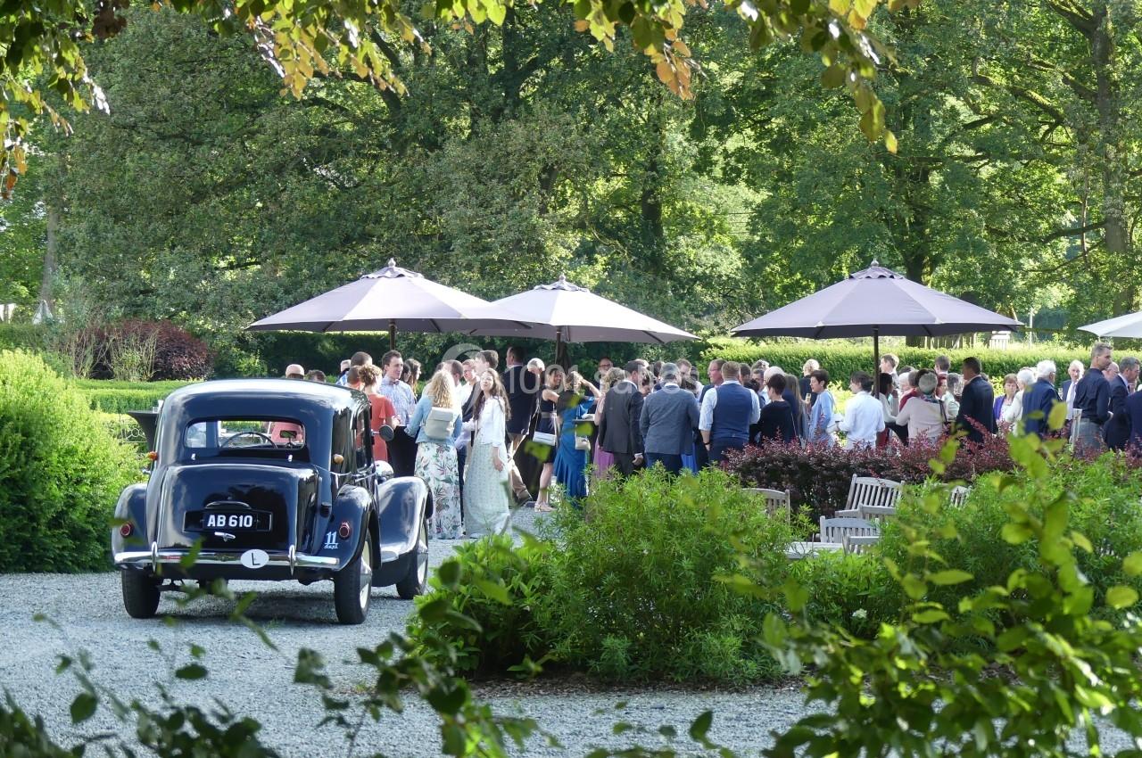 Un rassemblement de personnes dans un jardin, près de parasols, avec une voiture ancienne garée au premier plan.