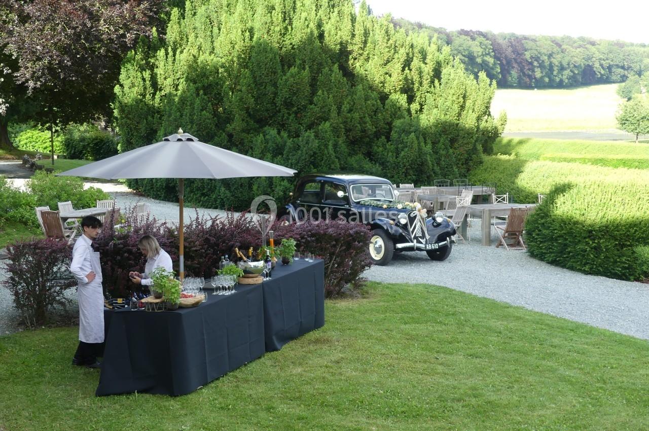 Deux serveurs préparent une table de buffet en extérieur, près d'une voiture ancienne et d'un jardin verdoyant.