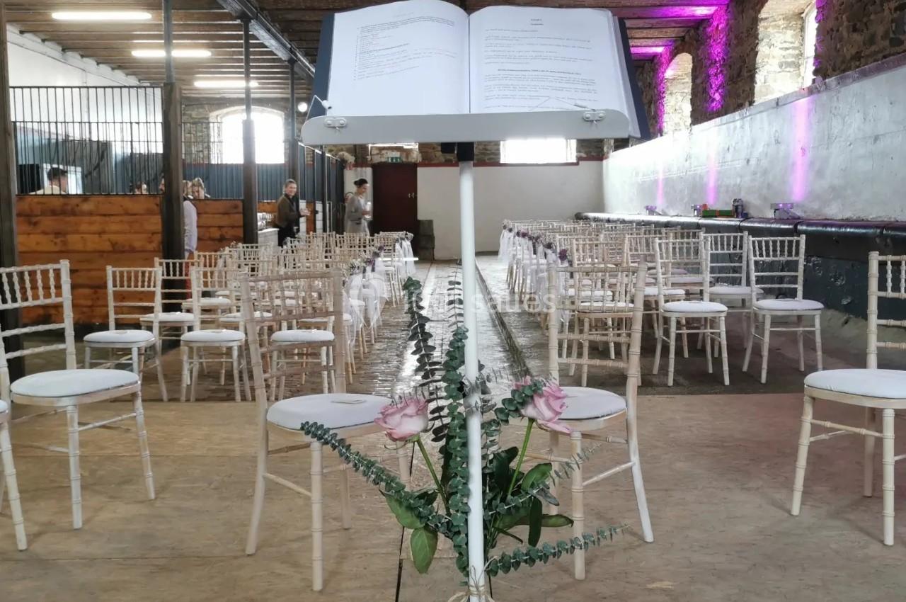 Salle de cérémonie avec des chaises blanches alignées et un pupitre décoré de fleurs au premier plan.