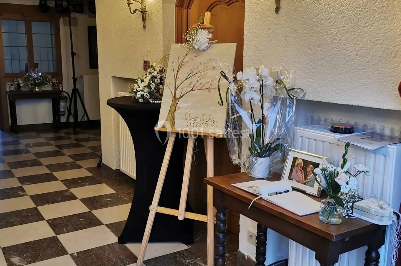 Chevalet avec tableau décoratif près d'une table ornée de fleurs et d'un livre d'or dans une salle lumineuse.